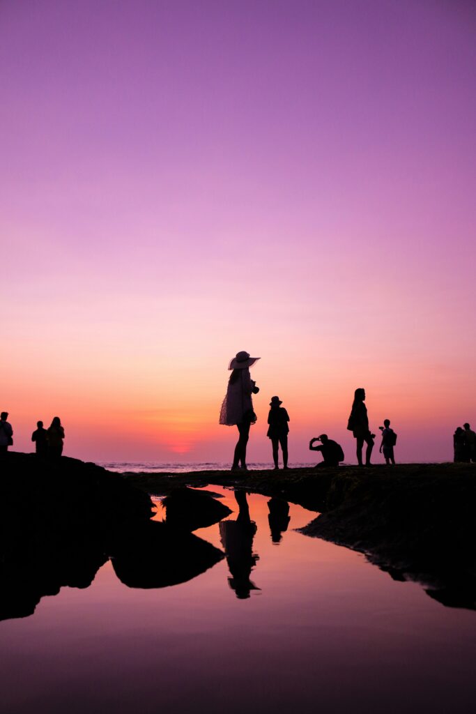 Silhouetted figures stand on the beach against a vibrant sunset, creating a serene and picturesque scene.