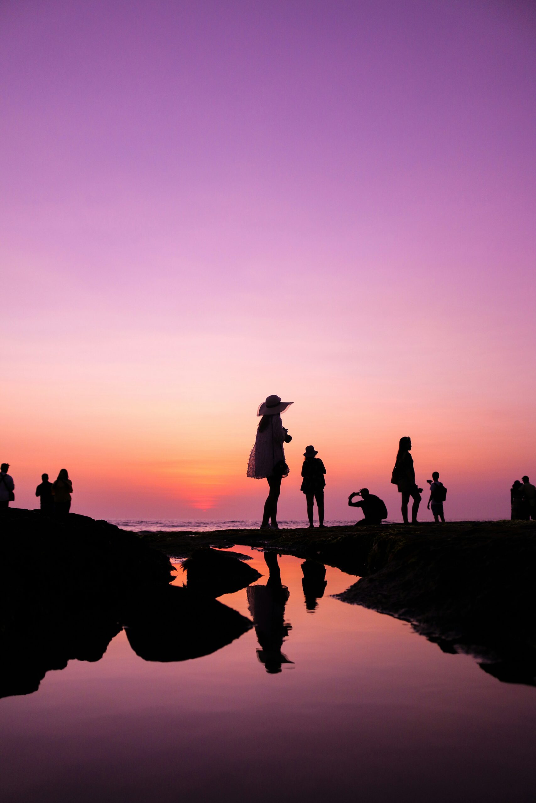 Silhouetted figures stand on the beach against a vibrant sunset, creating a serene and picturesque scene.